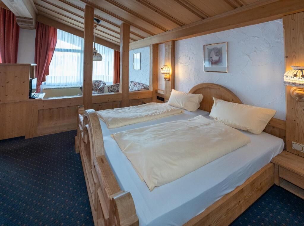 Cama doble con cabecera de madera, mesita de noche con lámpara y ventana con cortinas.