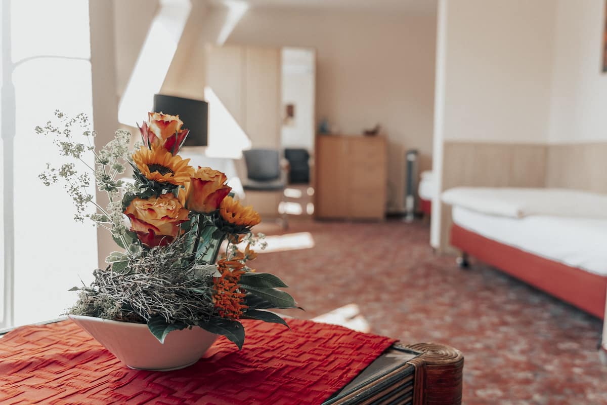 Une chambre d’hôtel avec un lit, une table en tissu rouge tissé et un vase de fleurs posé sur une table.