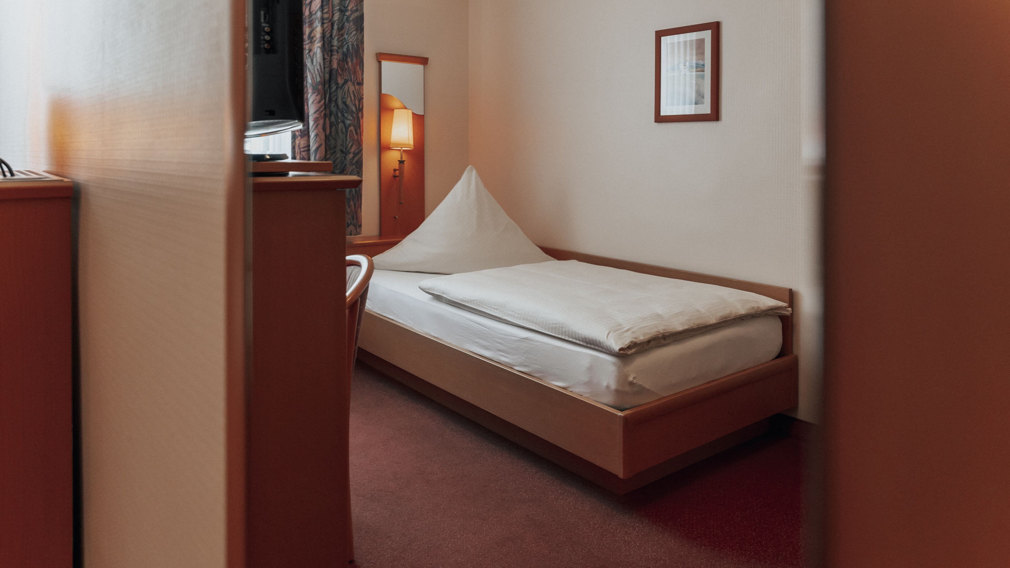A single bed with a wooden frame, a white mattress, and a white pillow. There is a small bedside table with a lamp, a framed picture on the wall, and a television in the corner.