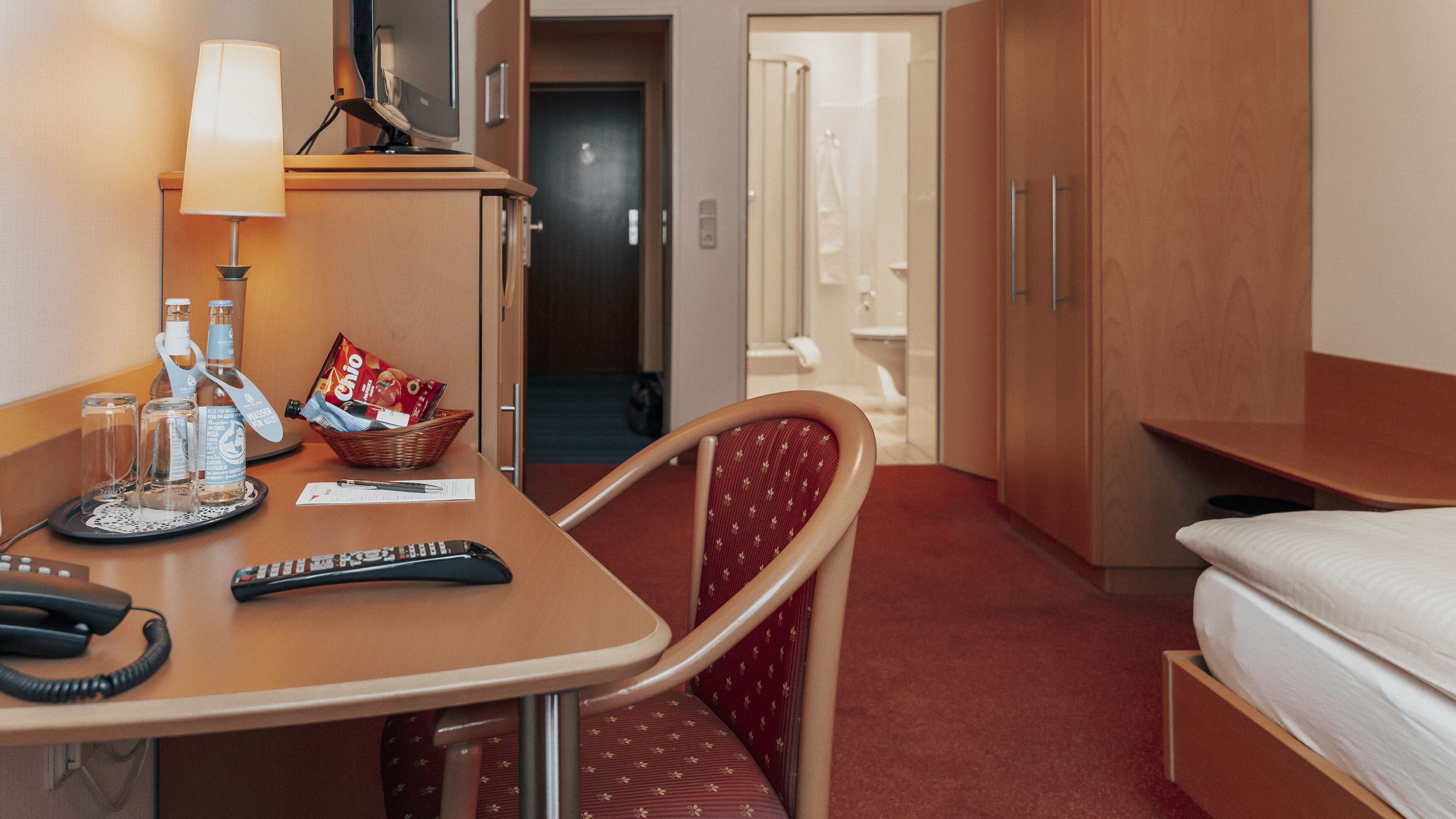 Ein Hotelzimmer mit Bett, Schreibtisch mit Lampe, Stuhl, einer Korb mit Snacks und einer Fernbedienung.