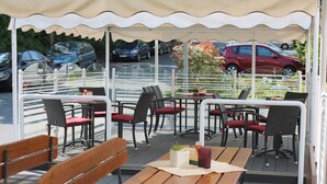 Un patio al aire libre con mesas y sillas de madera, un paraguas blanco y vistas a coches estacionados.