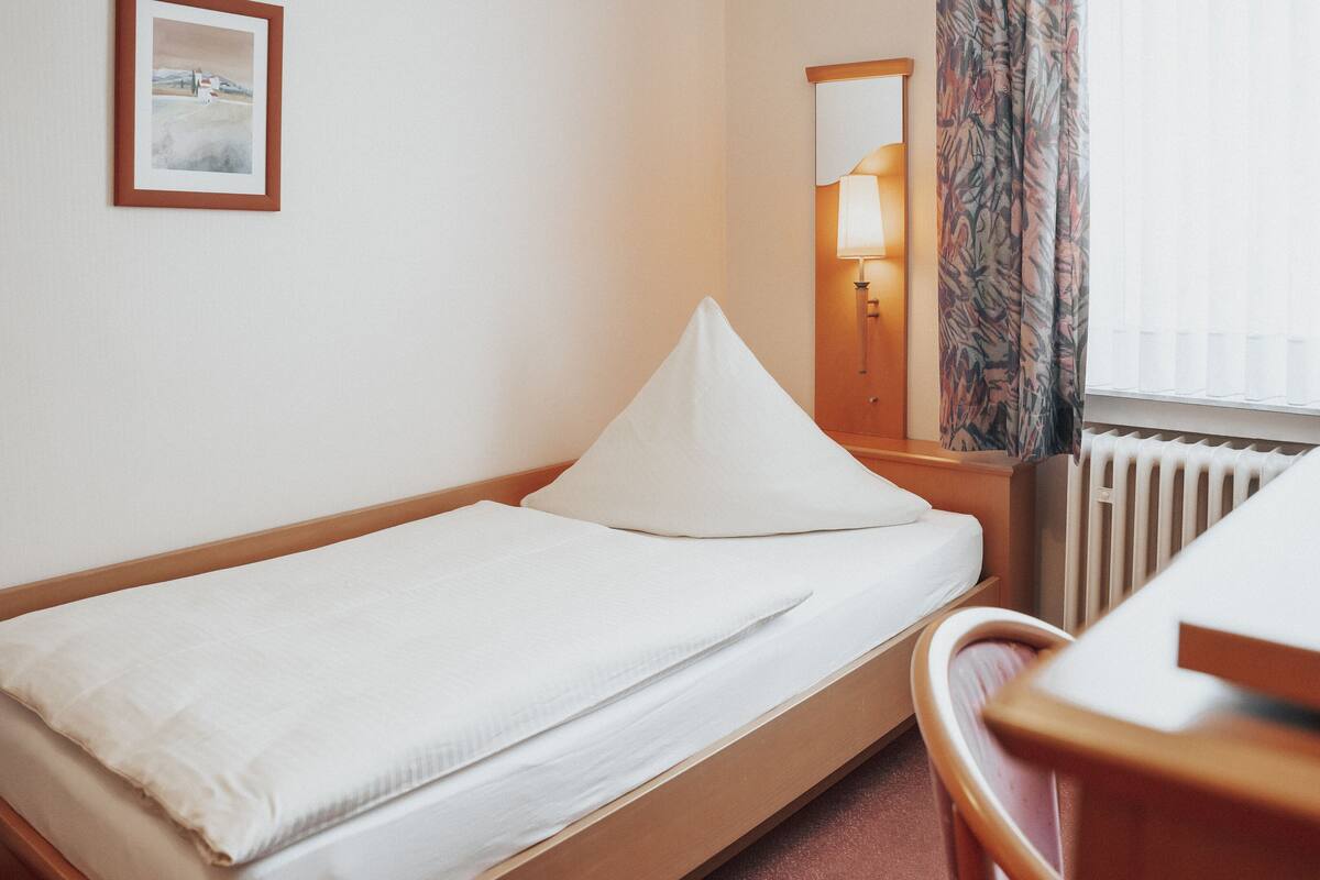 Un lit simple avec un matelas et un oreiller blancs, une tête de lit en bois, une petite table de chevet, une lampe, un miroir et un radiateur.