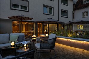 Un espacio al aire libre con mesas, sillas y sombrillas, con vistas a un restaurante con iluminación interior.
