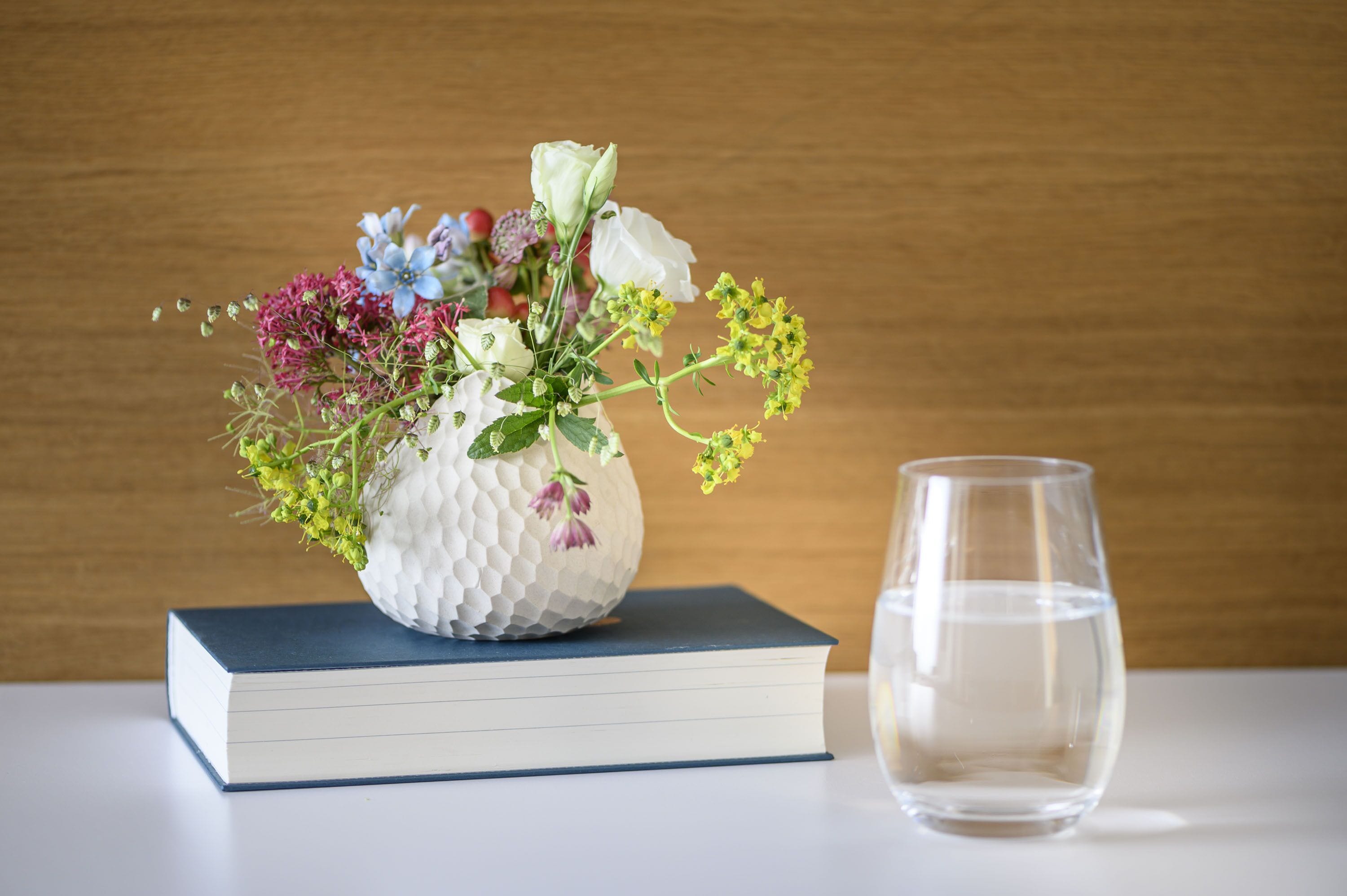 A white textured vase with flowers on a book, next to a clear glass.