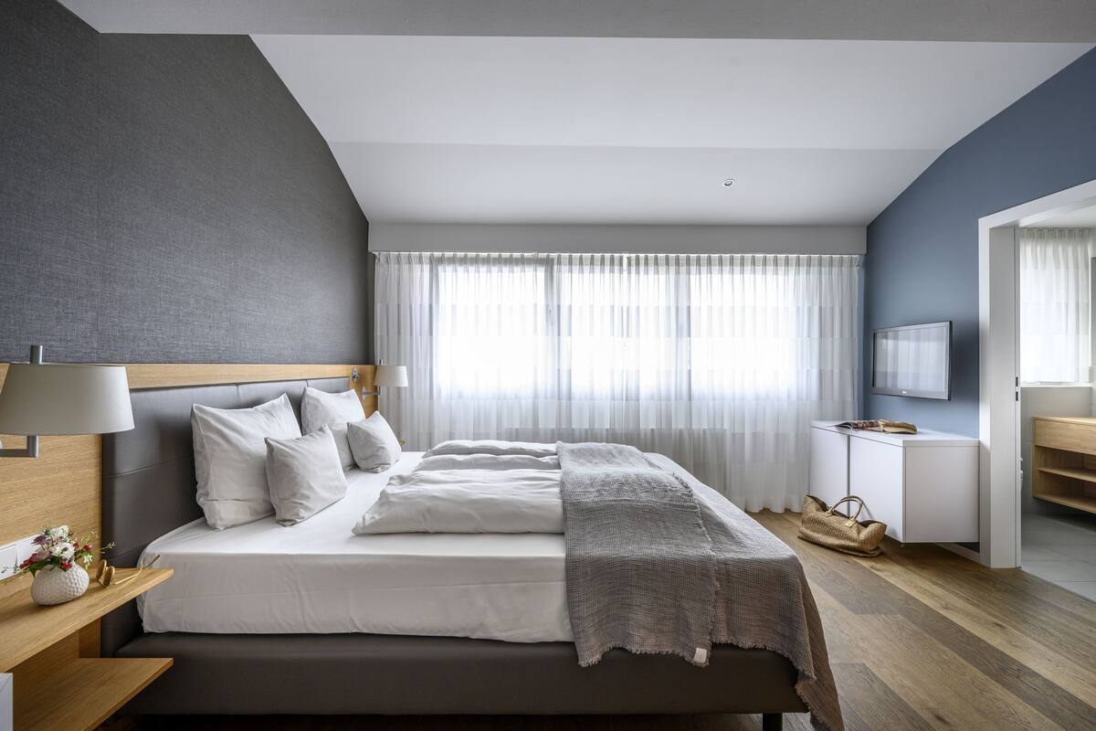 A modern bedroom with a large bed, a bedside table with a lamp, a bedside plant, and a view of a cityscape through sheer curtains.