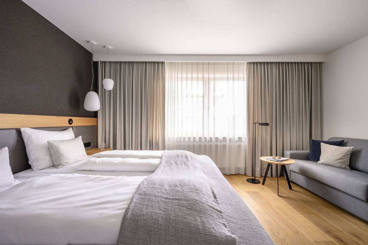 A modern hotel room with a large bed, a sofa, a small round table, and a large window with curtains.