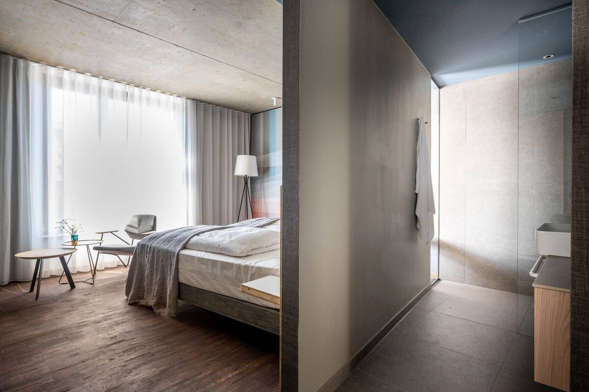 A modern hotel room with a bed, a desk, a chair, a lamp, and a view of the outside.