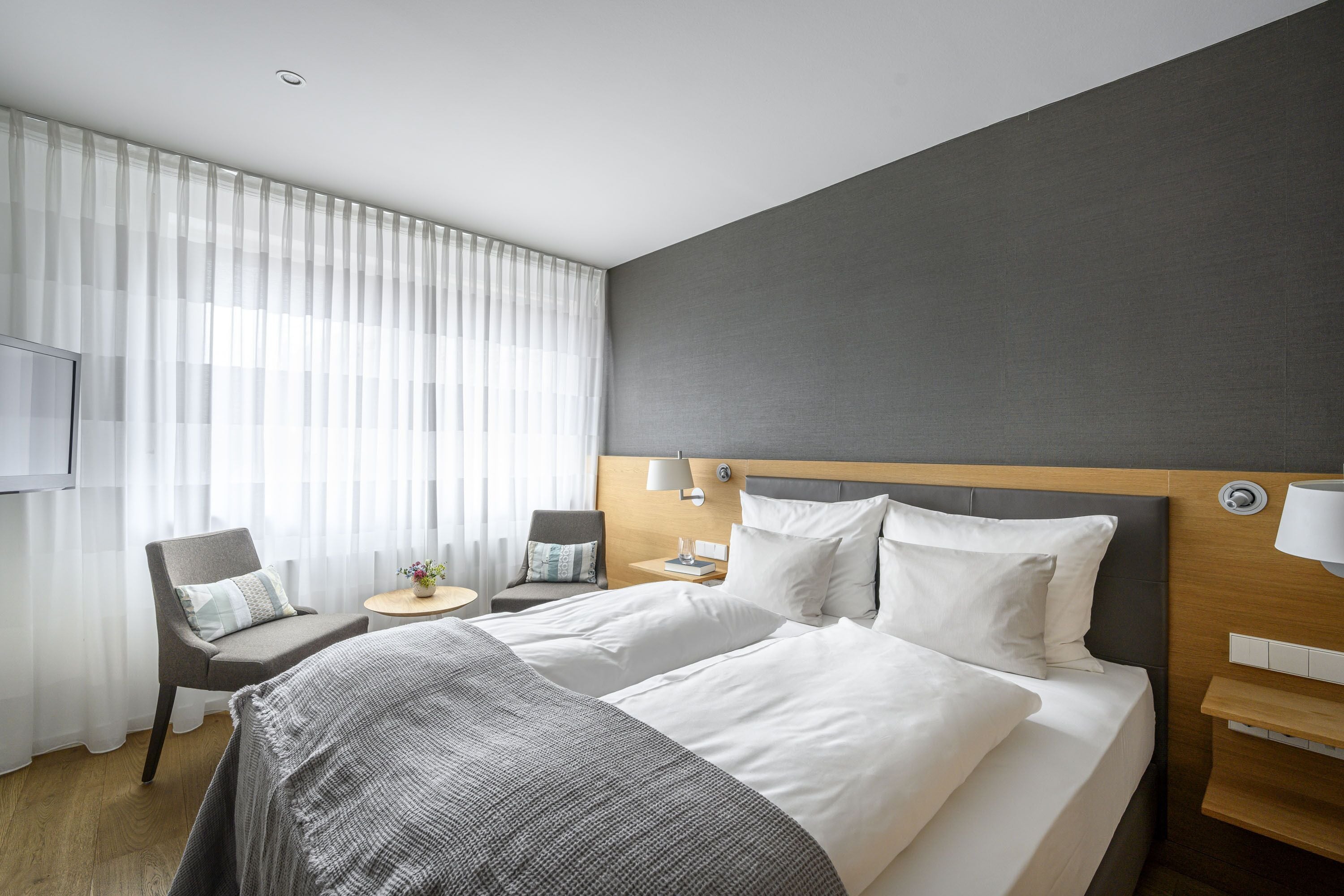 A hotel room with a large bed, a bedside table, a chair, a small table with a lamp, and a TV mounted on the wall.
