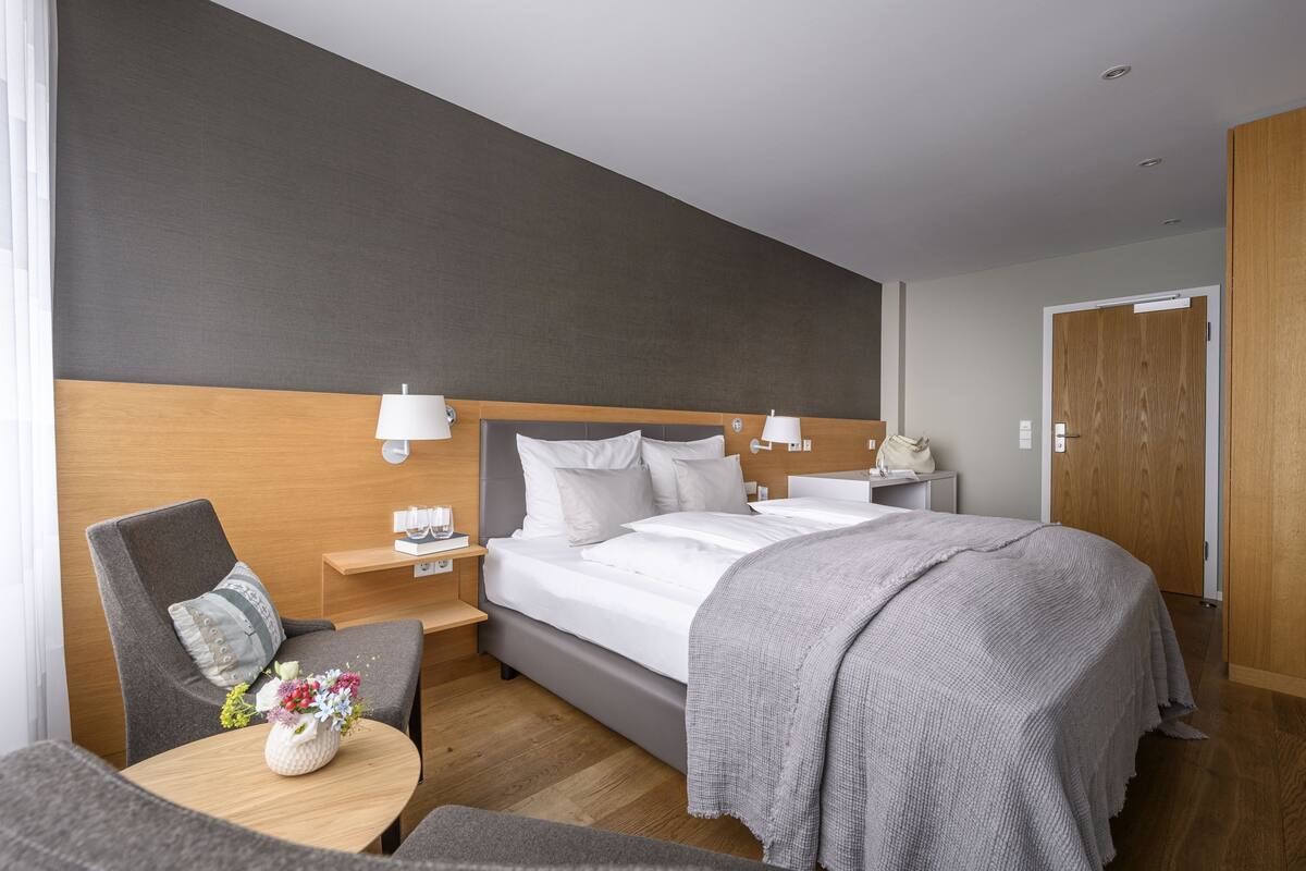 A hotel room with a bed, bedside tables, a chair, a small table with a vase, and a wooden wardrobe.