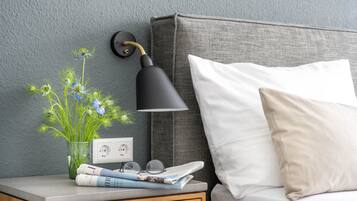 A neatly made bed with a white pillow, a bedside table with a lamp, a glass vase with flowers, and a power outlet.