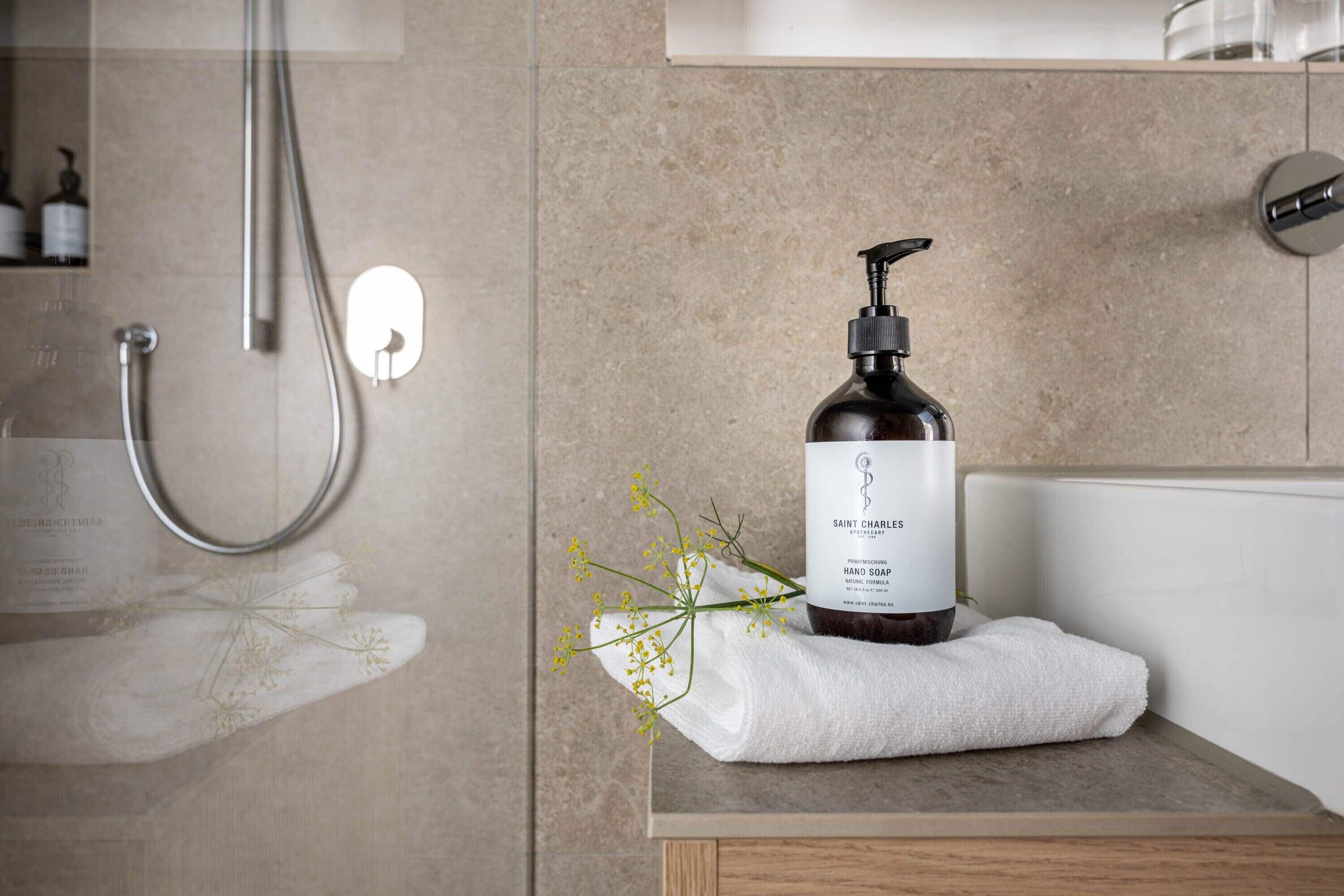 Ein Badezimmer mit Dusche, einer Flasche Saint Charles Handseife, einem weißen Handtuch und einer kleinen Pflanze.