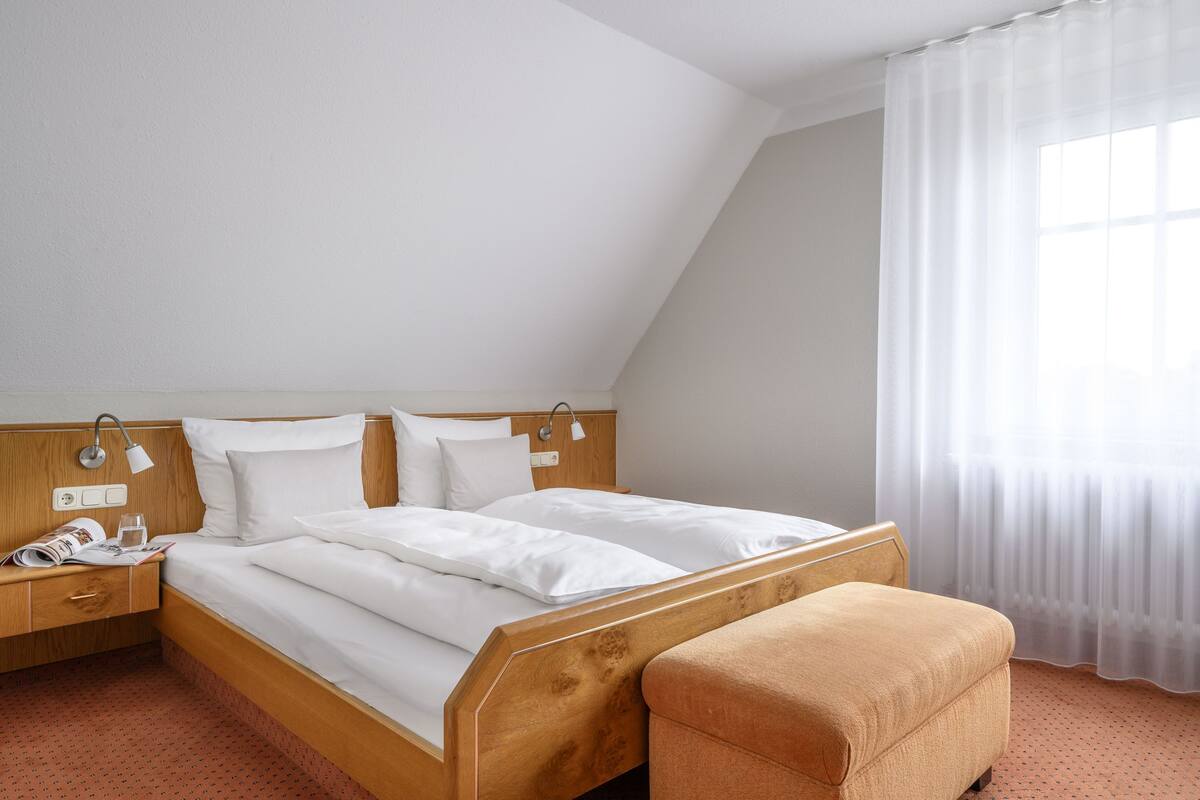 A hotel room with a wooden bed, a nightstand with a lamp, a window with sheer curtains, and a small wooden bench.