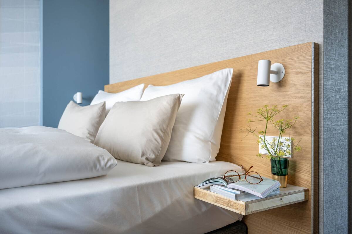 A neatly made bed with white linens and pillows, a bedside table with a book and glasses, and a wall-mounted light fixture.