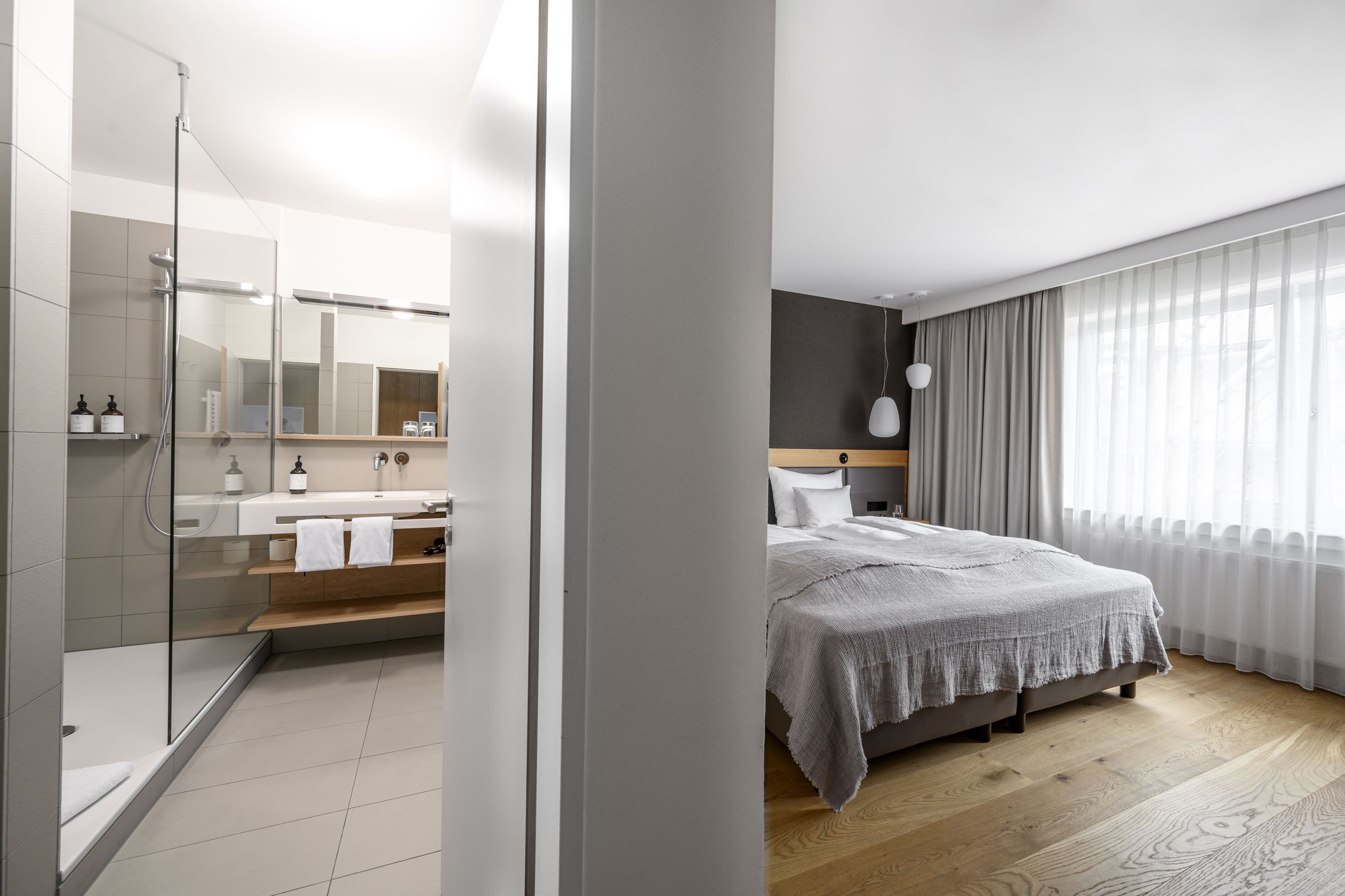 Habitación de hotel moderna con ducha, cama grande, tocador con espejo y vistas al exterior a través de cortinas traslúcidas.