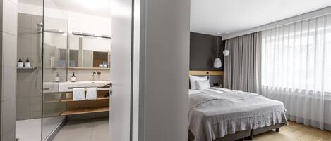 Habitación de hotel moderna con ducha, cama grande, tocador con espejo y vistas al exterior a través de cortinas traslúcidas.