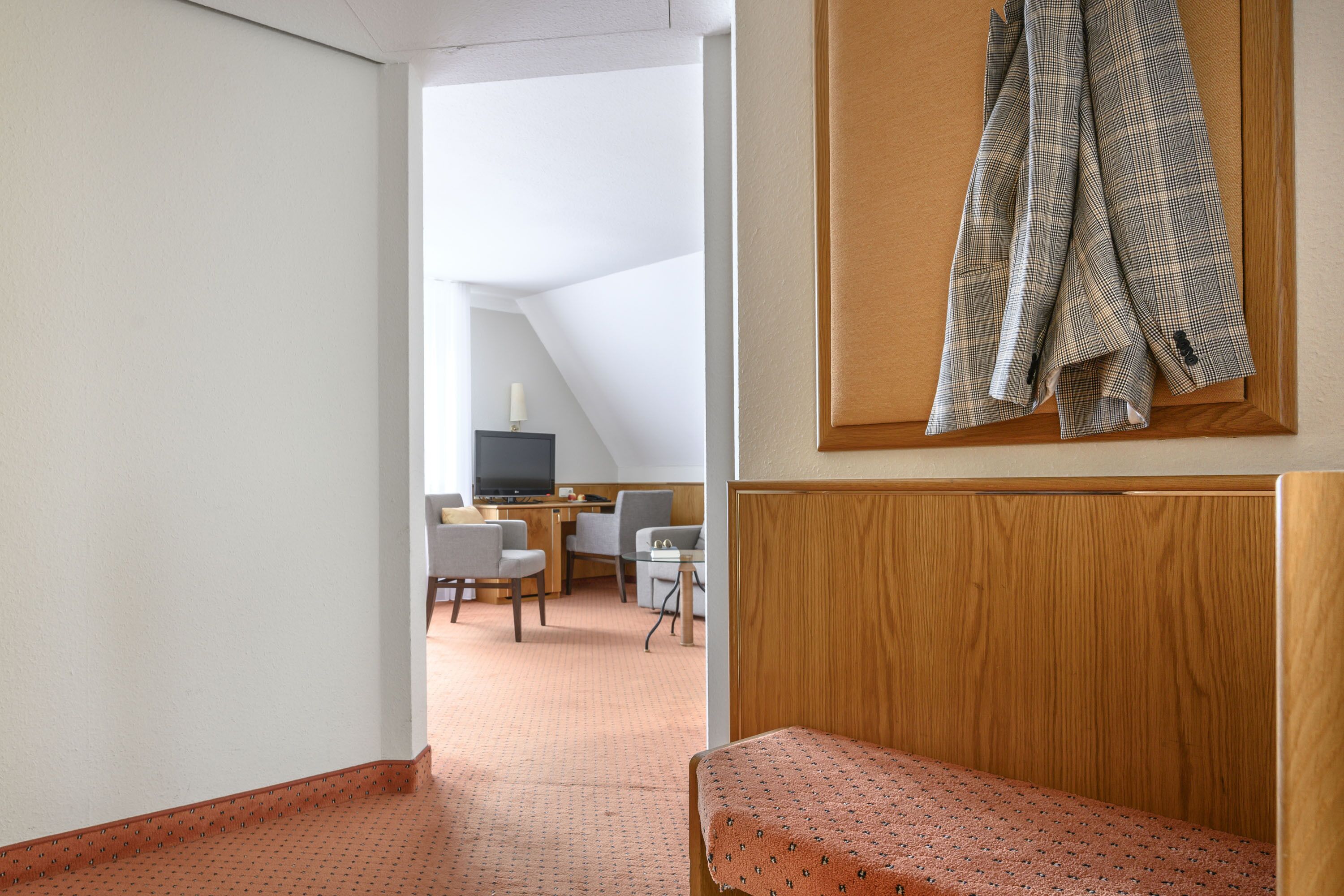 Quarto de hotel com um banco de madeira, uma escrivaninha com cadeiras e uma televisão.