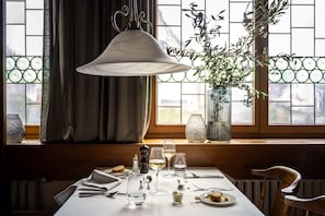 A dining table set with wine glasses, plates, and cutlery, a hanging light fixture, and a potted plant by a window with a view of a mountain. - Hotel Maier (Friedrichshafen)
