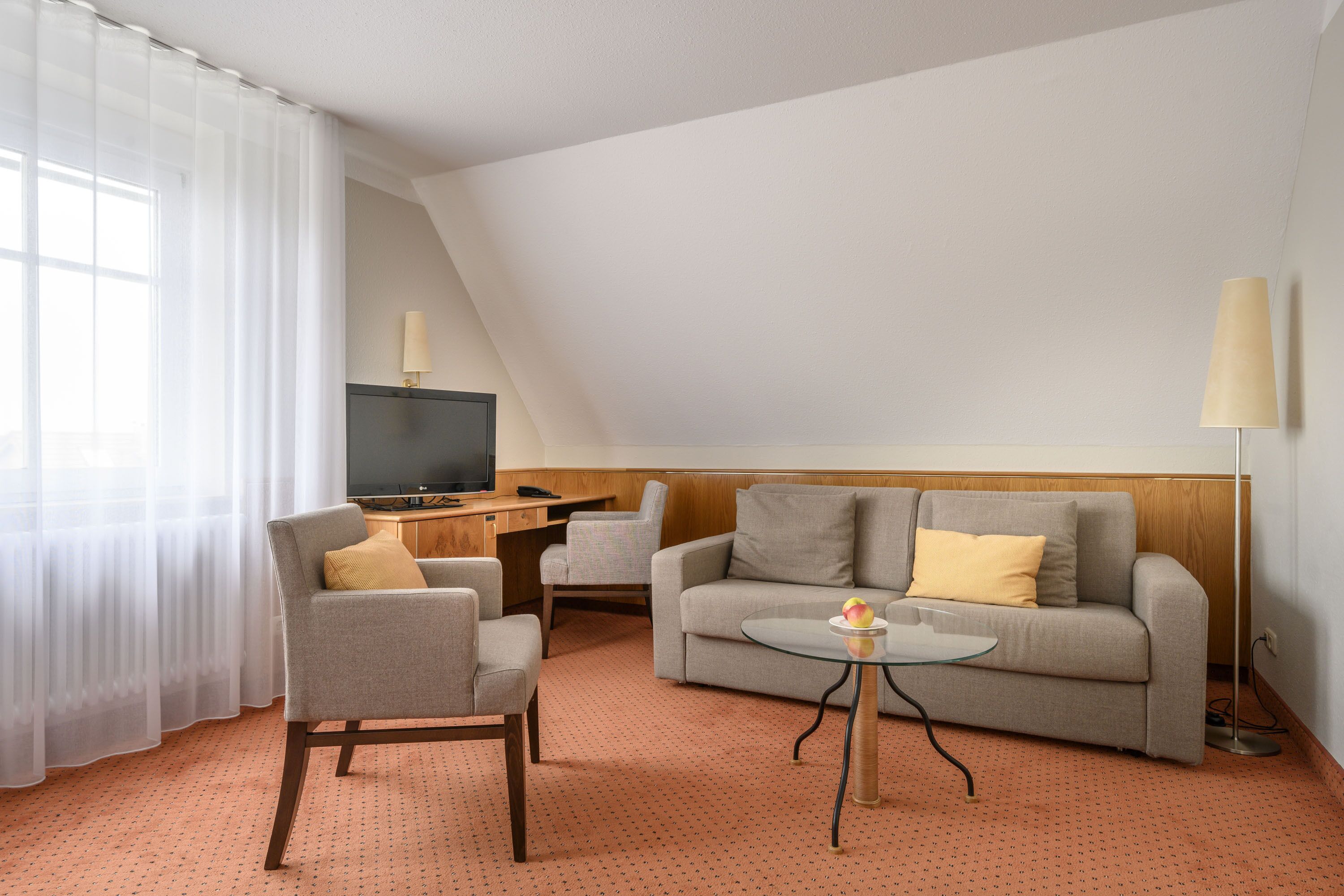 A hotel room with a sofa, armchair, and a small table with a lamp.
