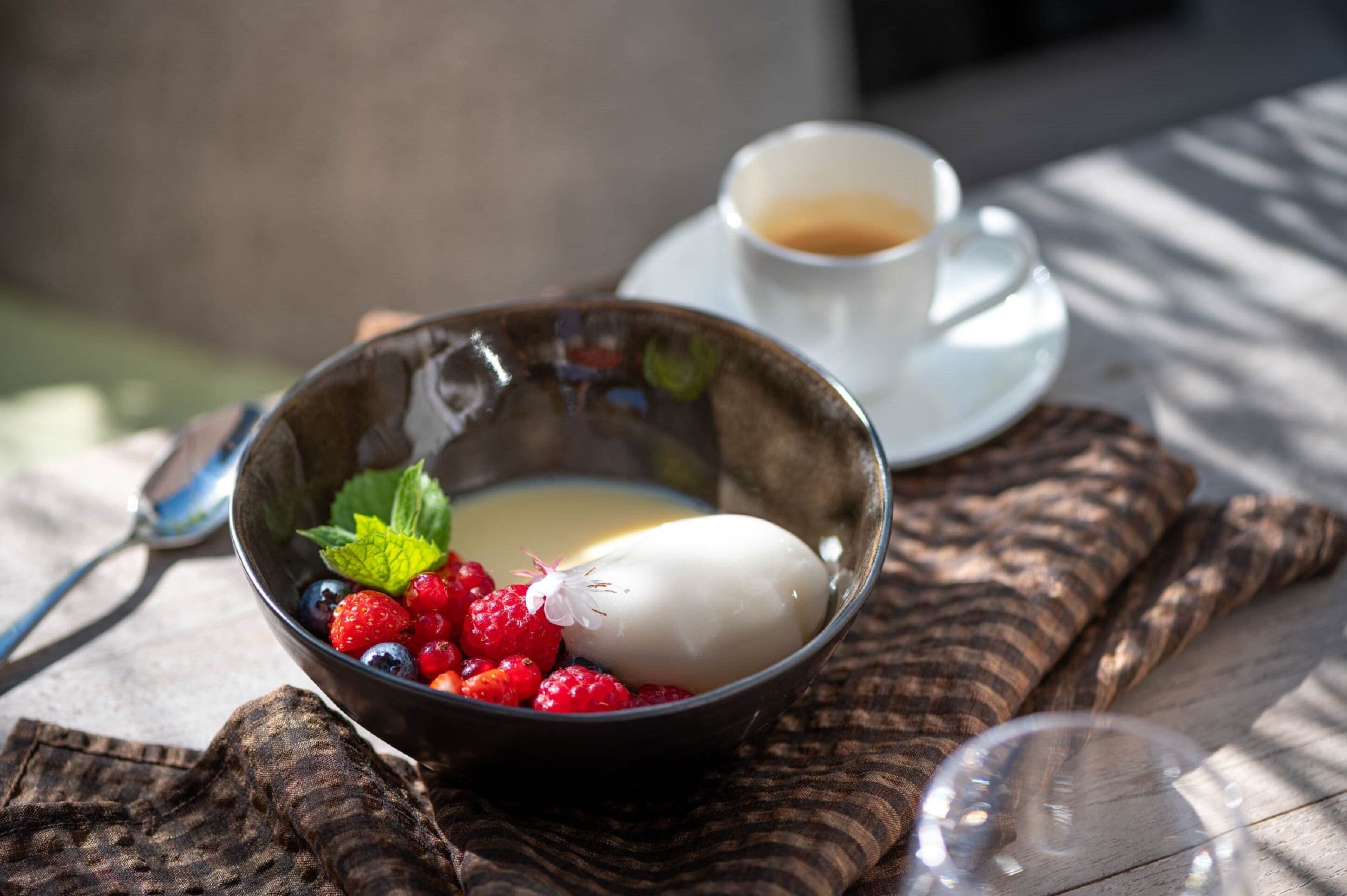 Un tazón de postre con bayas y una porción blanca, una taza de café y un vaso de agua sobre una mesa de madera.