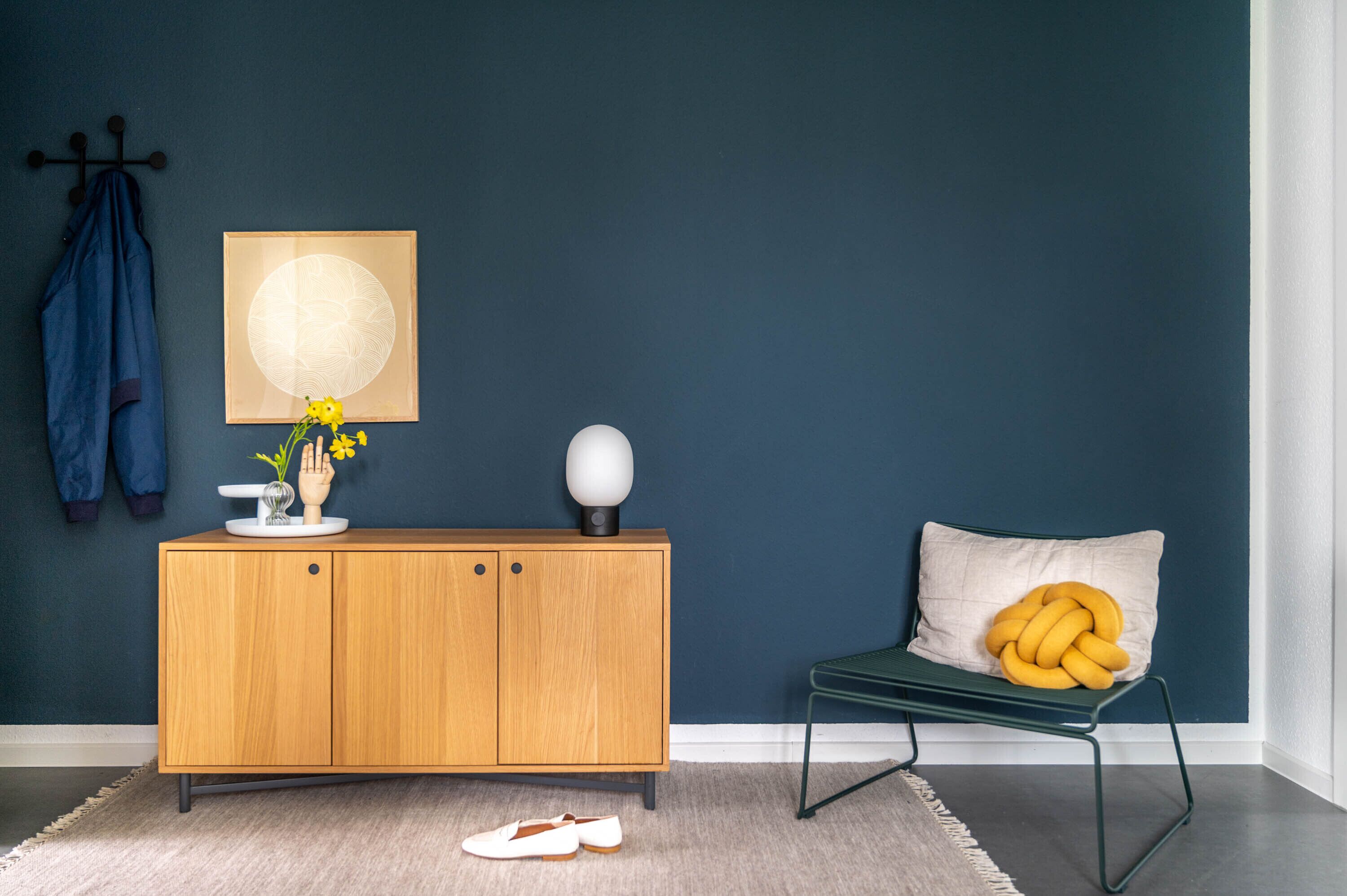 A room with a wooden cabinet, a chair with a yellow cushion, a coat rack with a blue jacket, and a wall-mounted artwork.
