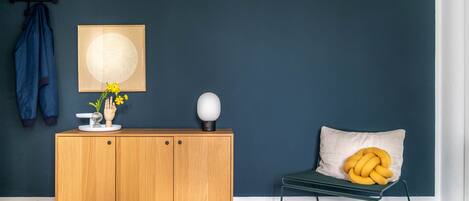 A room with a wooden cabinet, a chair with a yellow cushion, a coat rack with a blue jacket, and a wall-mounted artwork.