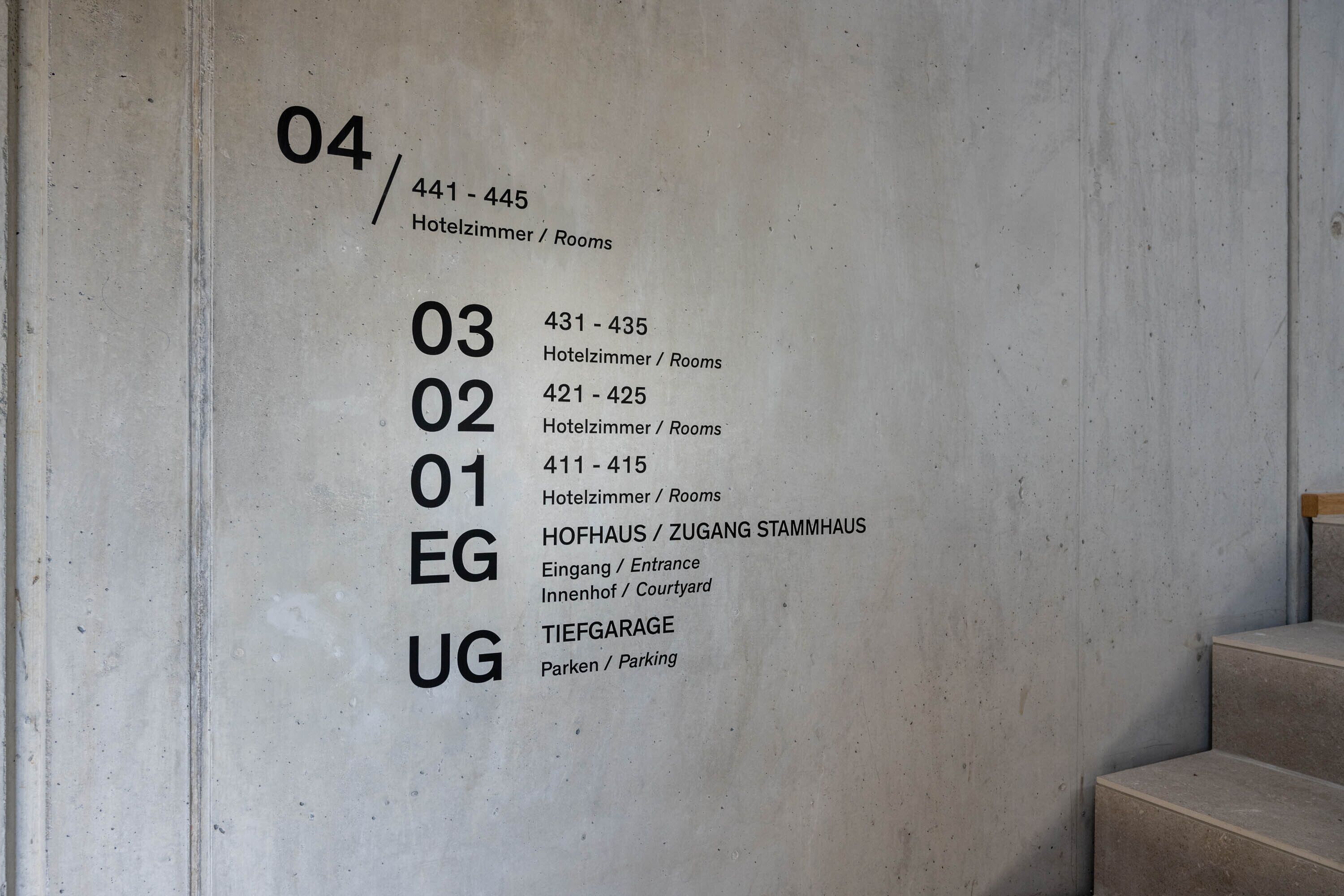 A wall with room numbers and corresponding descriptions: 04/441-445 (Hotelzimmer/Rooms), 03/431-435 (Hotelzimmer/Rooms), 02/421-425 (Hotelzimmer/Rooms), 01/411-415 (Hotelzimmer/Rooms), EG (Hofhaus/Zugang Stammhaus), UG (Tiefgarage).