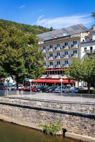 A hotel with multiple balconies, a stone wall, and parked cars along a river.