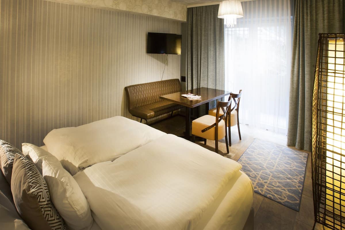 A hotel room with a large bed, a sofa, a dining table with chairs, a flat-screen TV mounted on the wall, and a window with curtains.