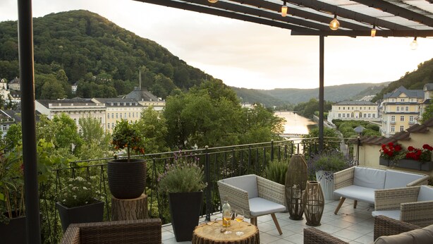 A terrace with wicker furniture, potted plants, and a view of a river and buildings. - Hotel Bad Emser Hof (Bad Ems)