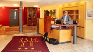 A hotel reception area with a reception desk, a red carpet with "BF SUPERIOR" lettering, a wooden cabinet with glass doors, and a red wall. - Hotel Bad Emser Hof (Bad Ems)