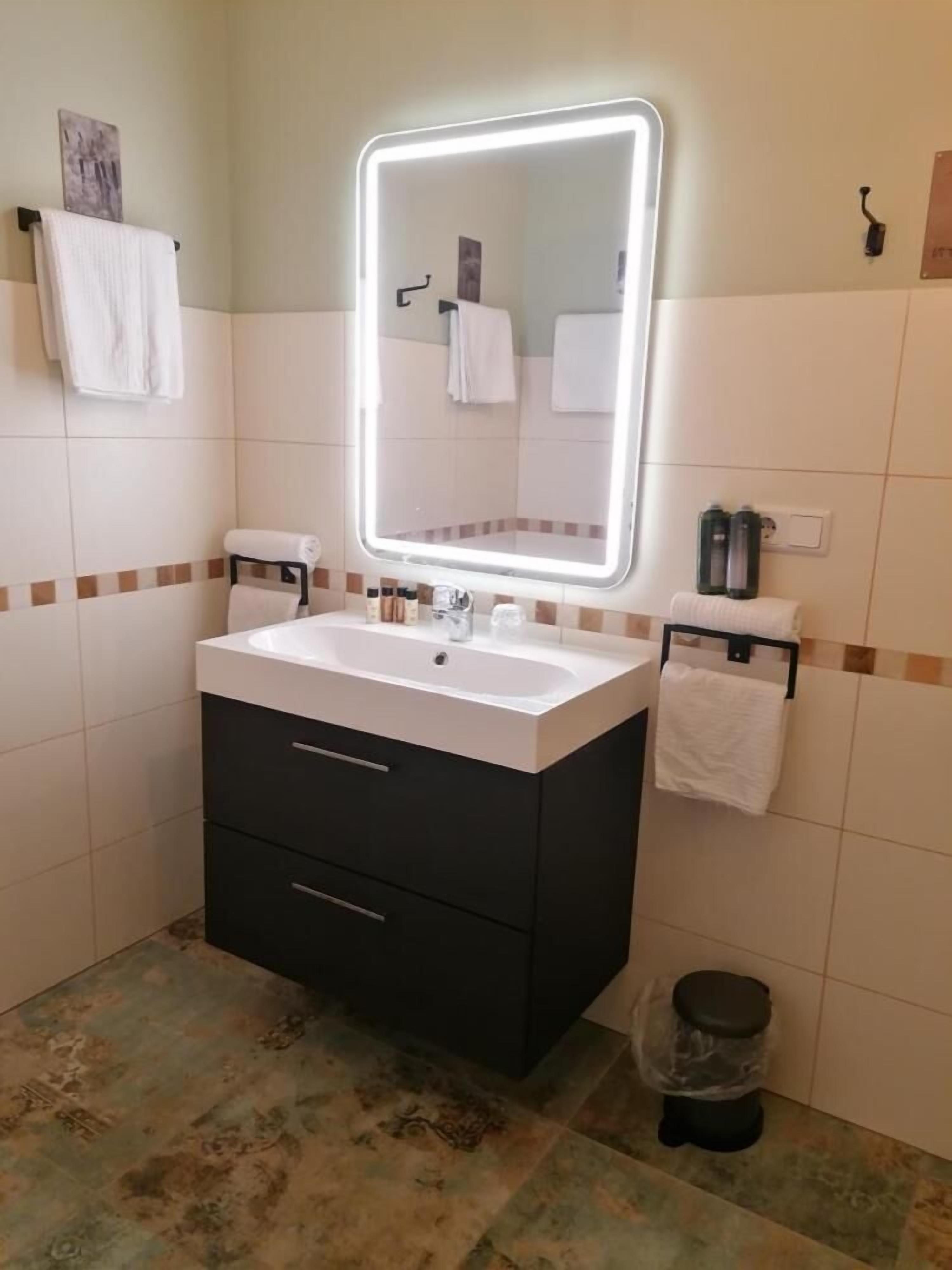 Un baño con lavamanos blanco, mueble de almacenamiento oscuro y un espejo con iluminación integrada. Hay toallas colgadas en la pared y un cubo de basura.