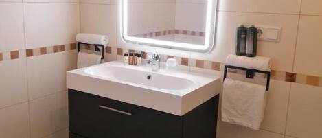 Un baño con lavamanos blanco, mueble de almacenamiento oscuro y un espejo con iluminación integrada. Hay toallas colgadas en la pared y un cubo de basura.