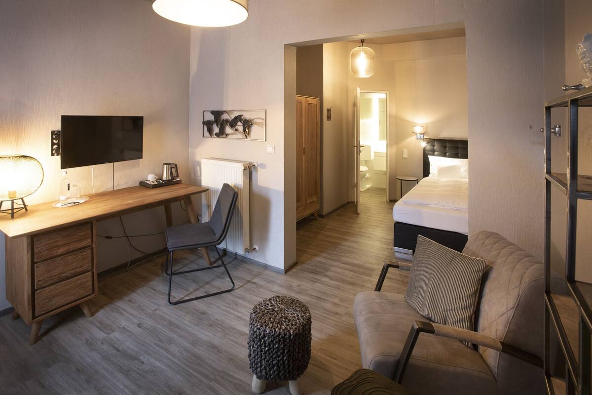 A hotel room with a wooden desk, a chair, a bed with pillows, a small ottoman, and a television.