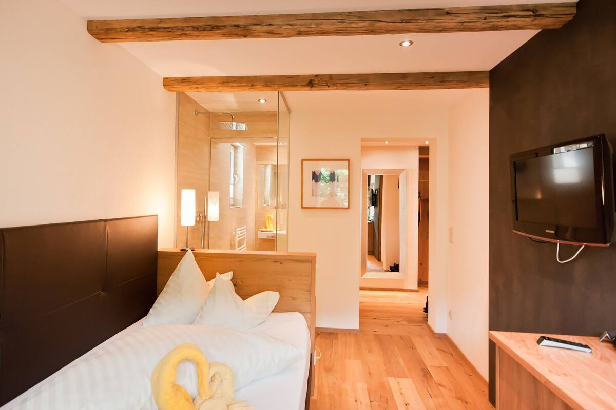 Habitación de hotel con techo de madera, cama con ropa blanca, televisor montado en la pared y mesita de noche de madera.
