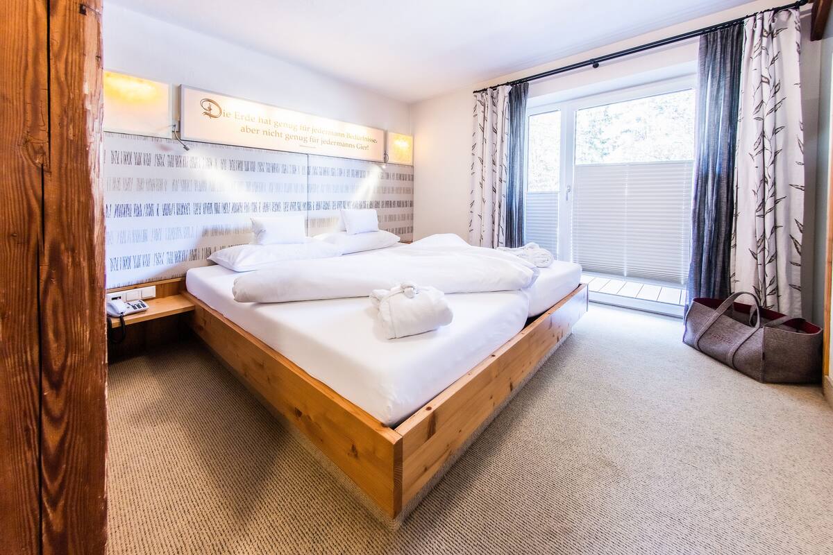 Una cama bien hecha con sábanas blancas y una toalla, cabecera de madera, mesita de noche con teléfono y una ventana con cortinas.