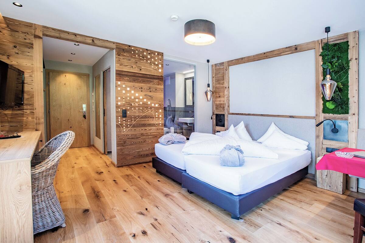 Una habitación de hotel moderna con suelo de madera, cama con ropa blanca, una silla de mimbre, un escritorio y una mesita con mantel rosa.