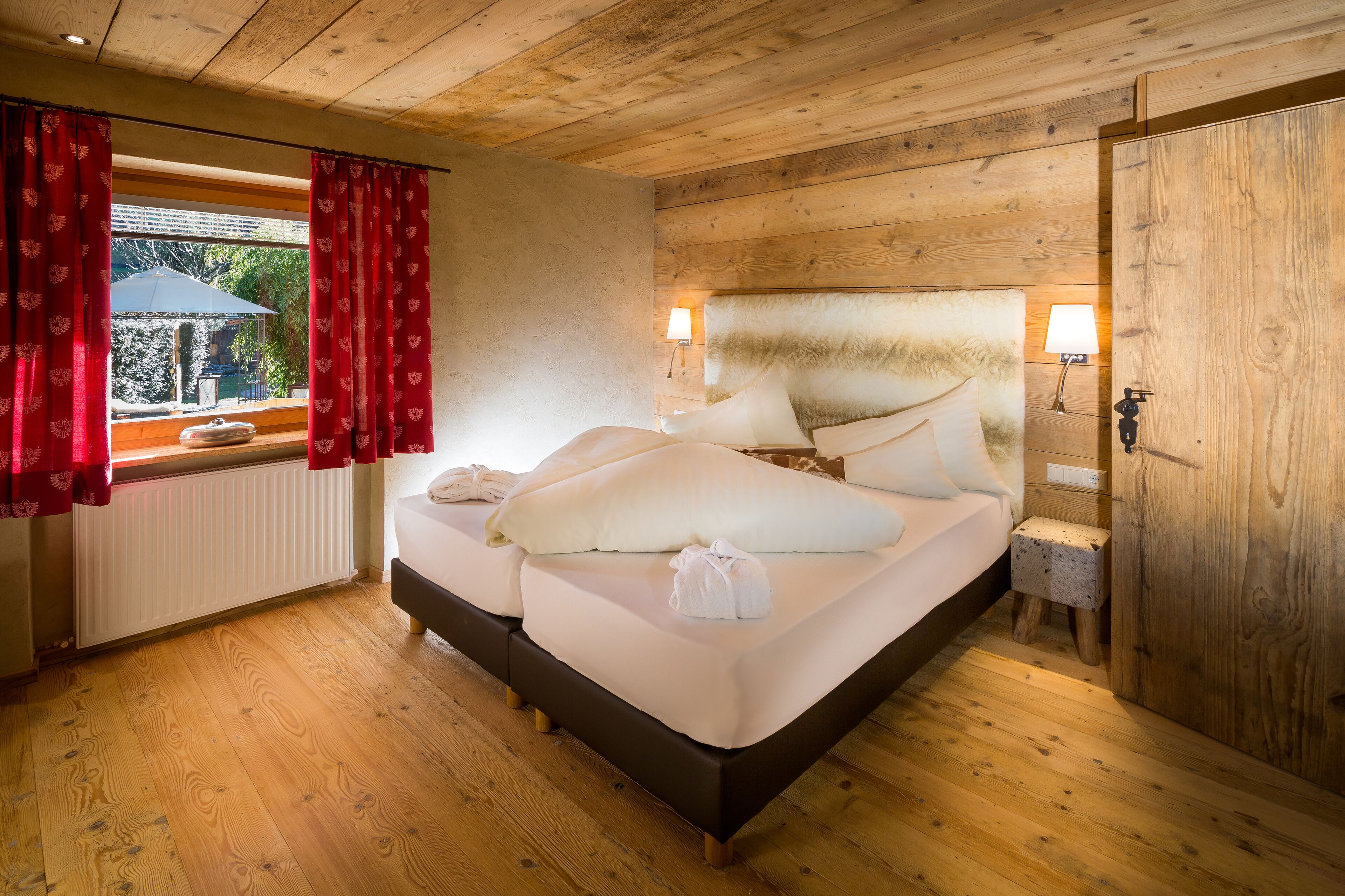 A wooden-paneled room with a bed, a window with red curtains, a lamp, and a radiator.