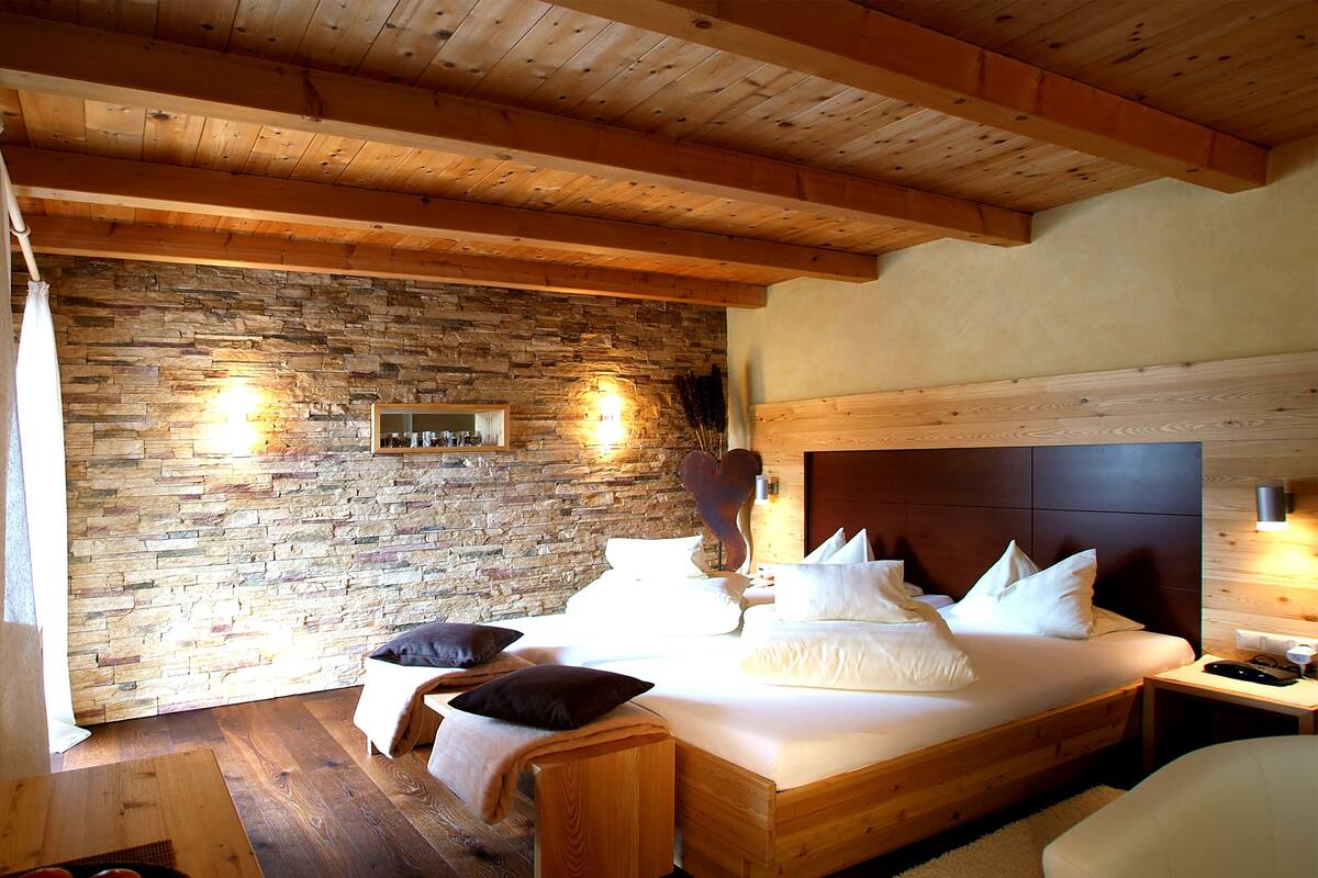 Un dormitorio con pared de piedra, vigas de madera en el techo, una cama con ropa de cama blanca y una mesita de noche con lámpara.