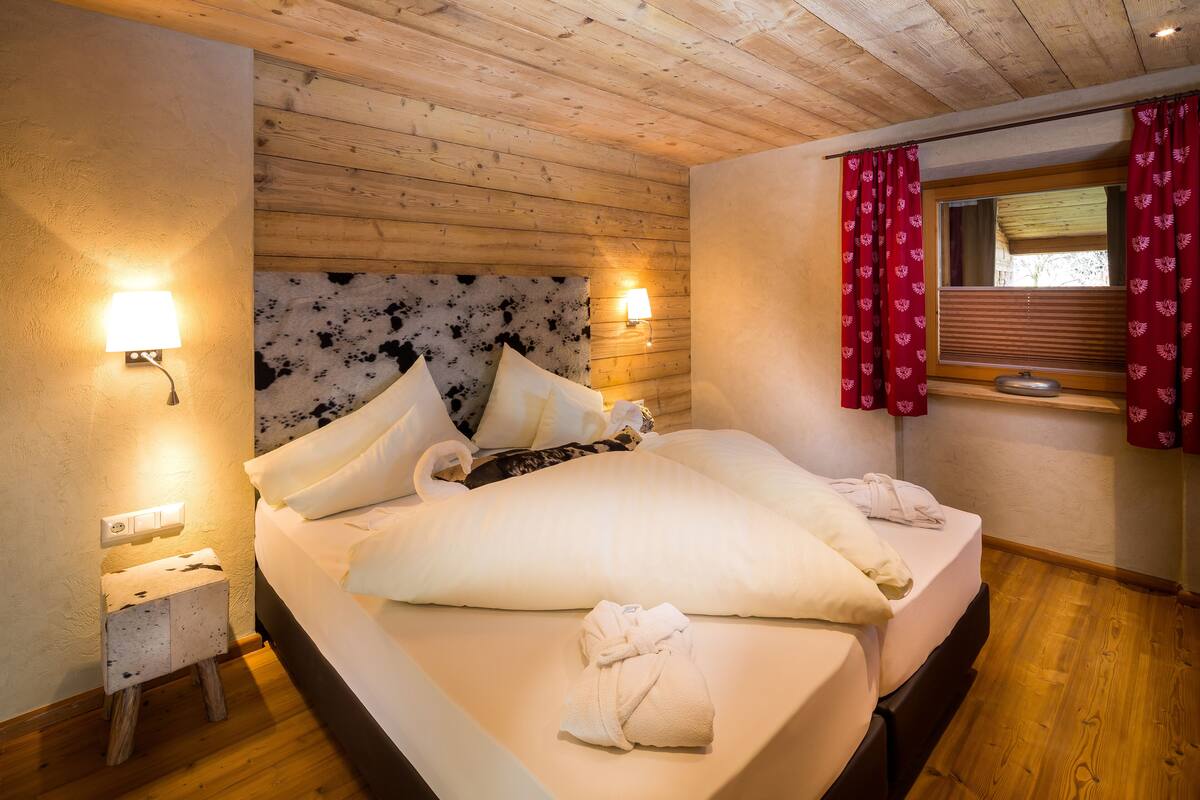 Cama doble con ropa de cama blanca, dos lámparas de noche, cabecera con estampado blanco y negro, y ventana con cortinas rojas.
