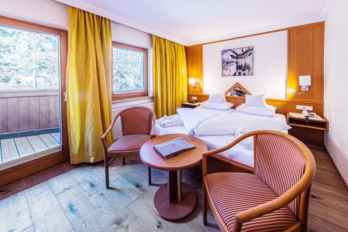 Habitación de hotel con cama, dos sillones, una mesita, ventana con cortinas y una lámpara de pared.