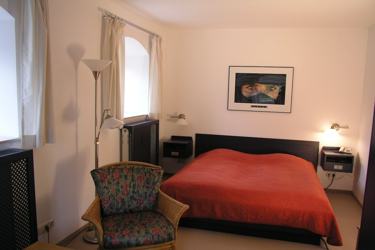 Ein Hotelzimmer mit einem Bett, einem Sessel, einem Schreibtisch, einer Lampe, einer Heizung und einem gerahmten Bild an der Wand.
