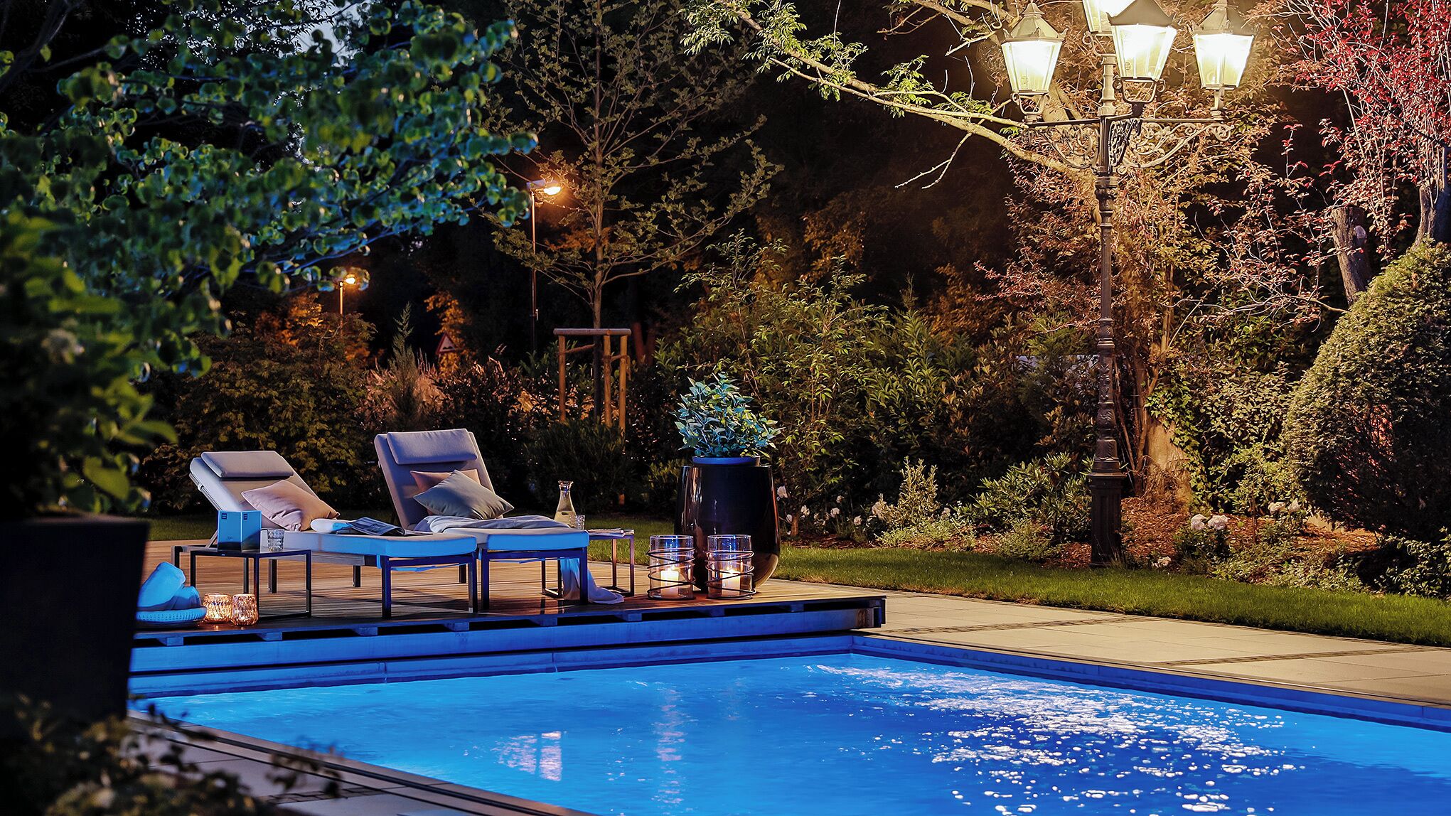 A swimming pool with lounge chairs and a table, surrounded by trees and a lamp post.