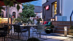 An outdoor seating area with tables and chairs, a potted plant, and a building with a red sign in the background. - Zeller Hotel & Restaurant (Kahl)