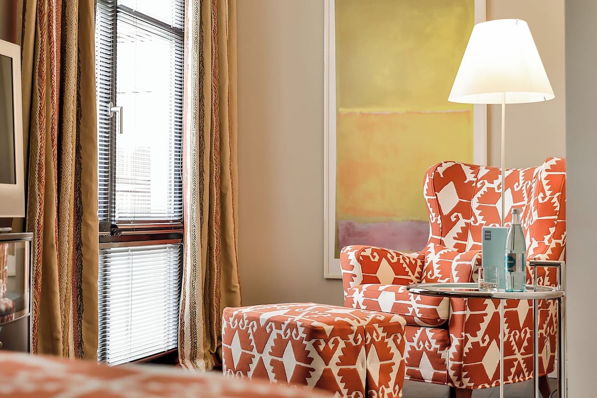 A hotel room with a television, a patterned armchair, a side table with a bottle, and a window with blinds.