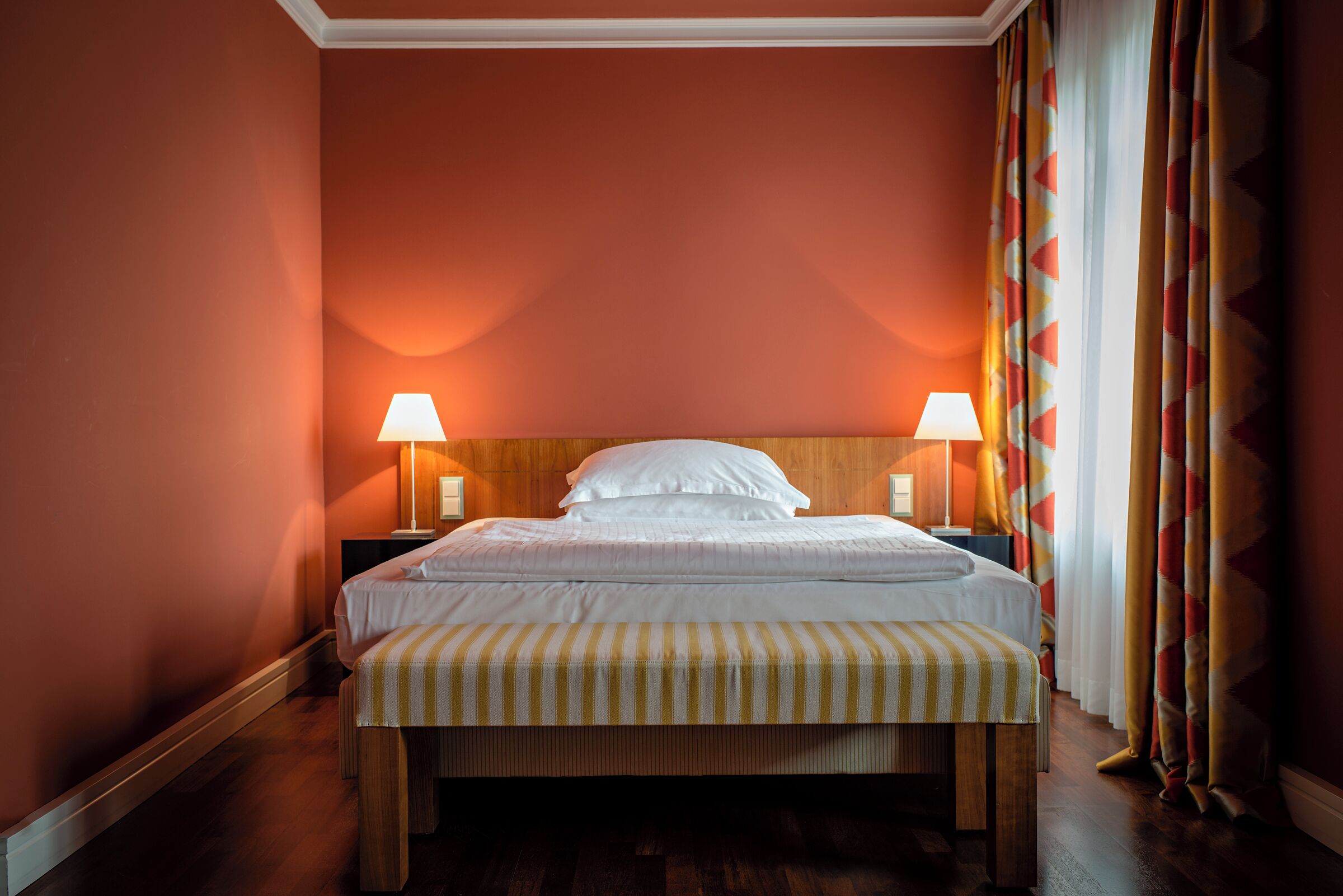 A hotel room with a large bed, two bedside lamps, a wooden headboard, and a striped bench.