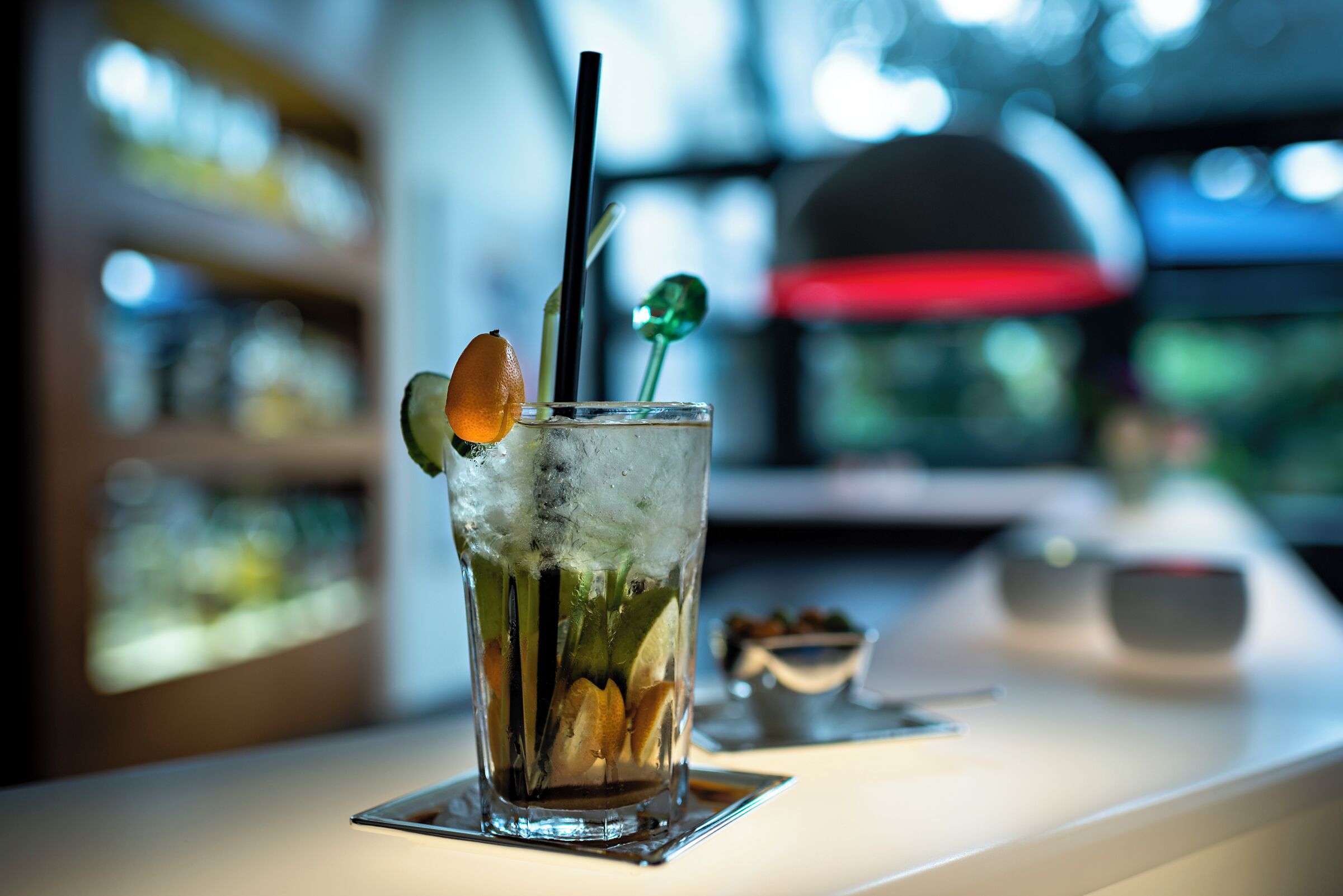 A cocktail with a straw and garnish on a coaster, with a blurred background of shelves and a red umbrella.