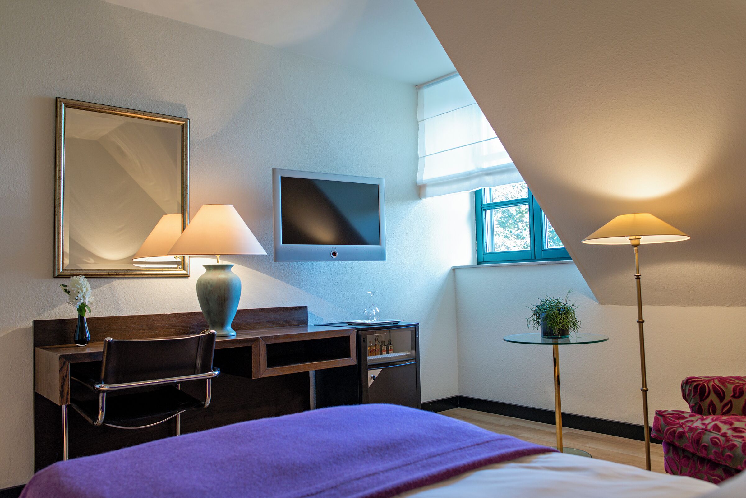 A hotel room with a bed, a desk with a chair, a TV, a lamp, a small table with a plant, and a window.