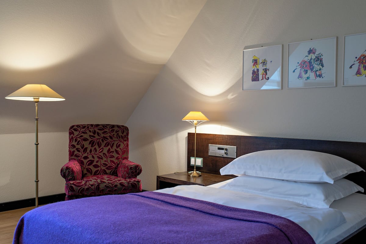 A hotel room with a bed, two bedside lamps, a chair, and three framed artworks on the wall.