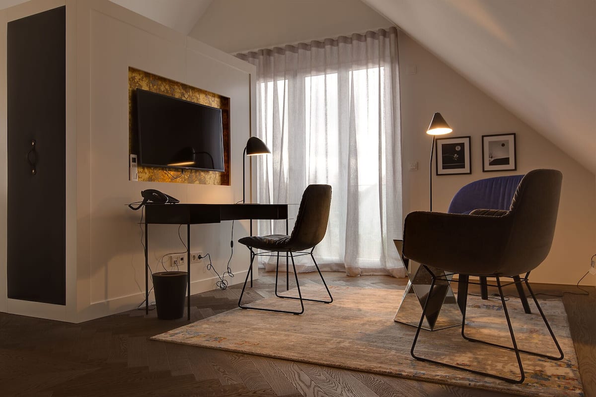 A modern living room with a flat-screen TV, a desk with a phone, two chairs, a large window with sheer curtains, and a floor lamp.
