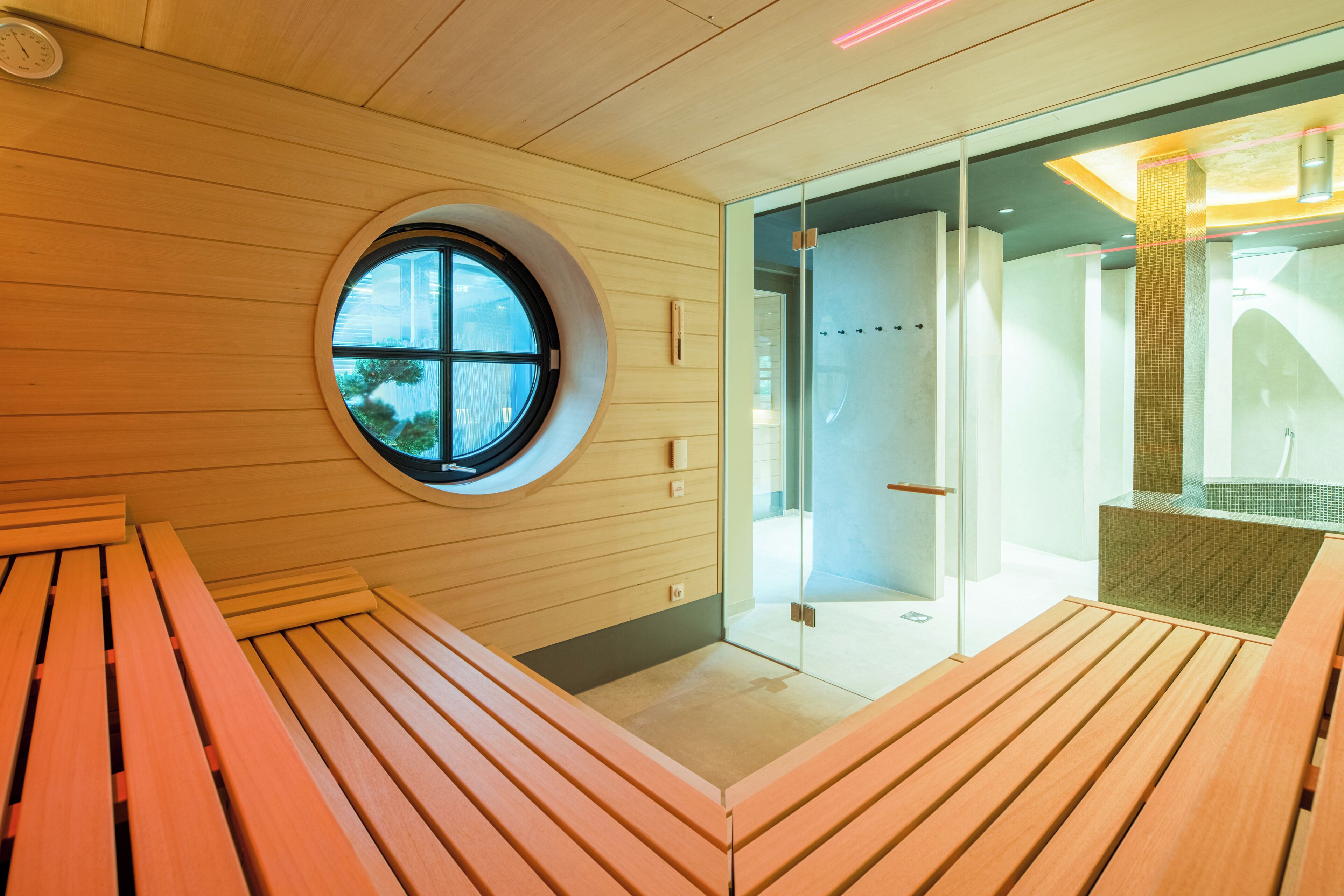 Un baño moderno con un banco de madera, un ventanal redondo y un área de ducha con mampara de vidrio.
